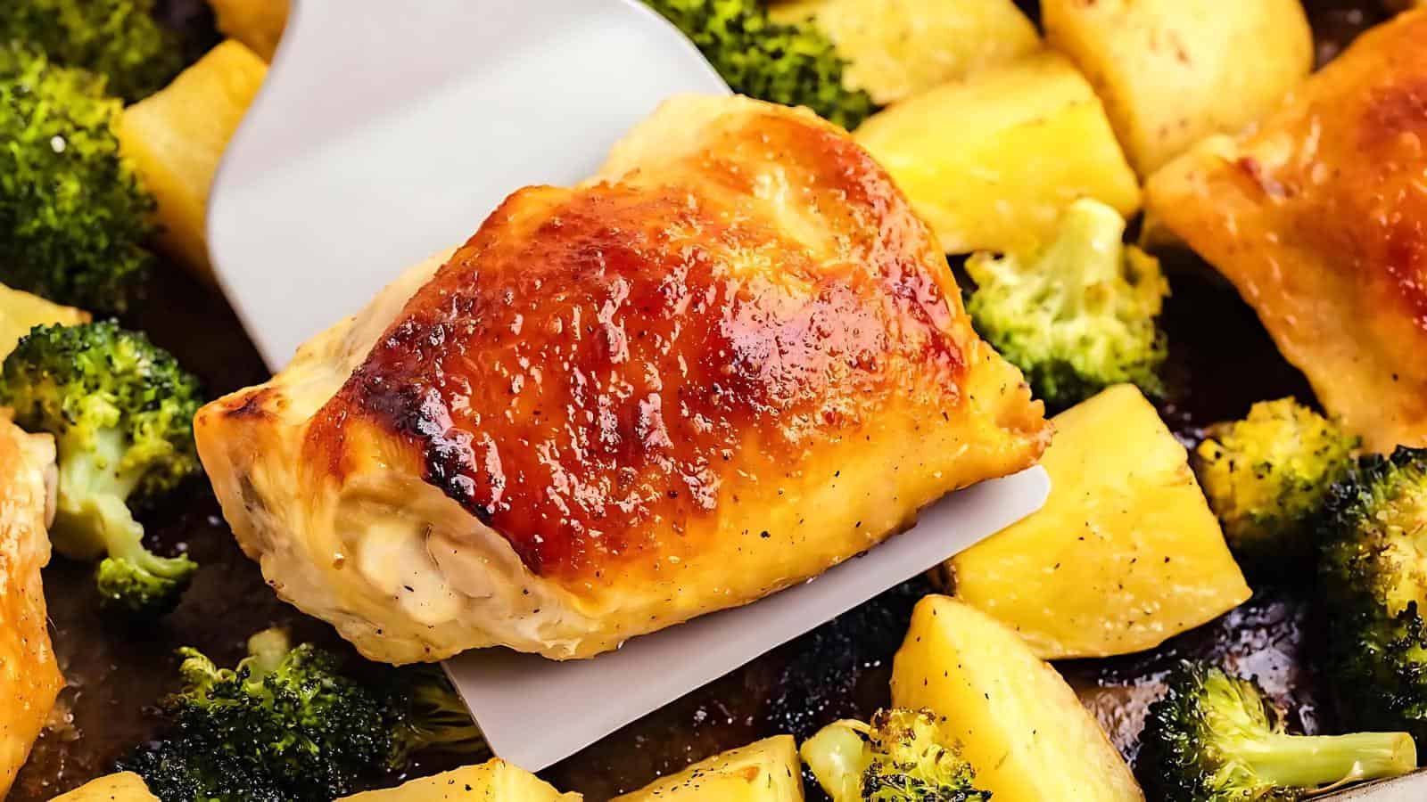 Roasted chicken thigh with golden skin and pineapple chunks on a spatula, surrounded by roasted broccoli and pineapple pieces.