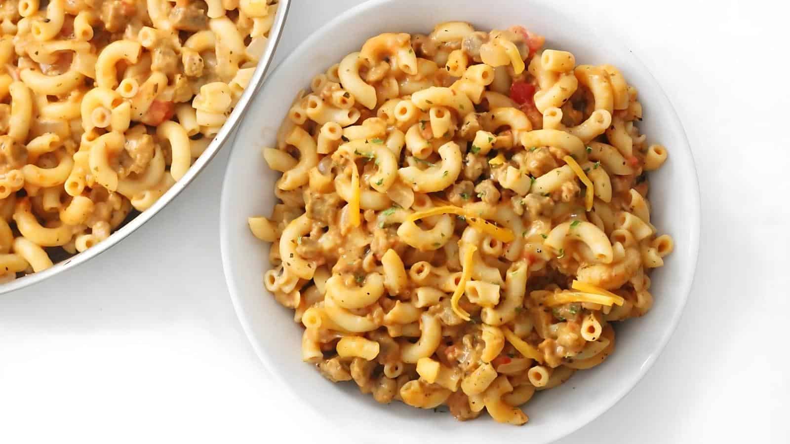 A bowl of creamy macaroni and cheese with ground meat and herbs, placed next to a partially visible pan of the same dish.