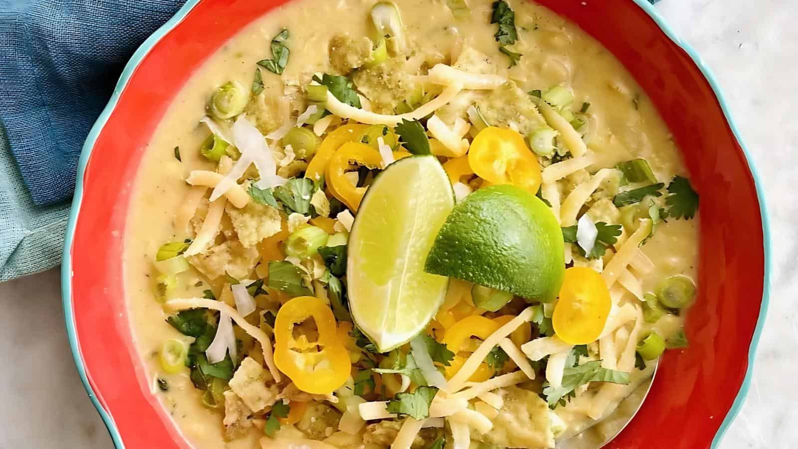 A bowl of soup topped with shredded cheese, tortilla chips, sliced yellow peppers, cilantro, and two lime wedges.