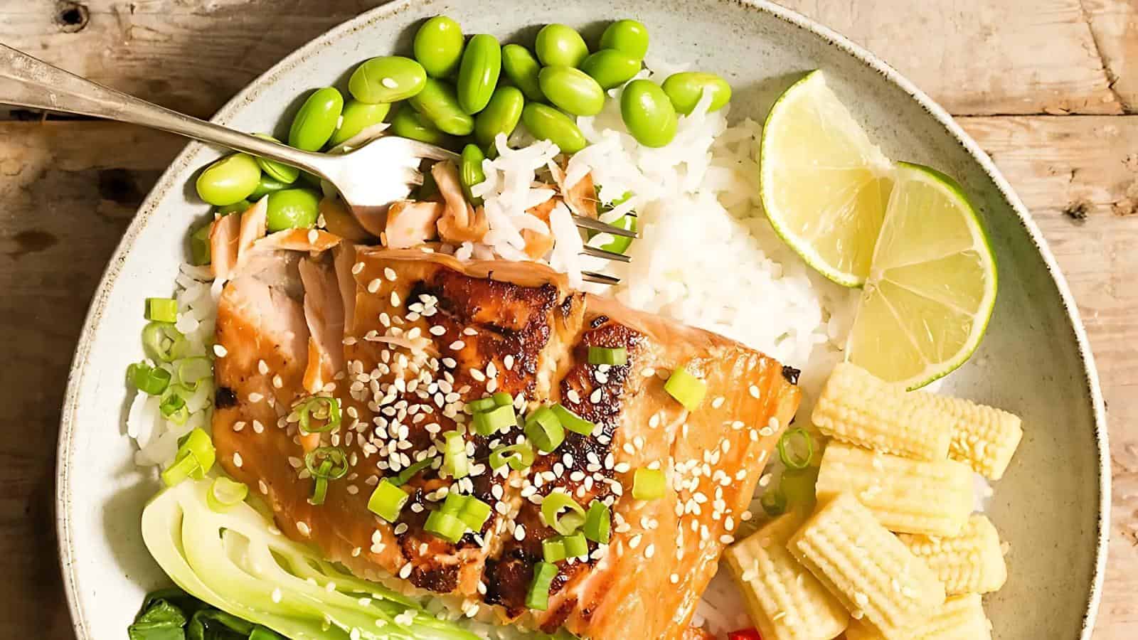 A bowl with rice, grilled salmon topped with sesame seeds and green onions, edamame, lime wedges, baby corn, and bok choy on a wooden surface.