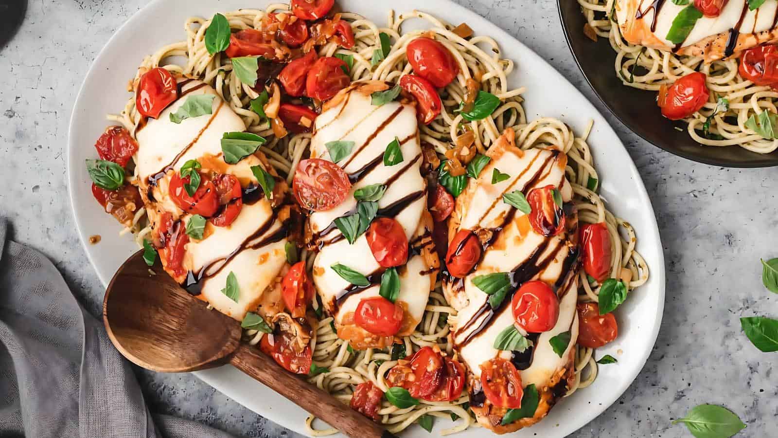Plate of spaghetti topped with grilled chicken, cherry tomatoes, basil, and balsamic glaze. Wooden spoon on the side.