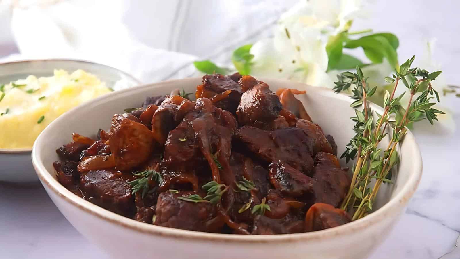 A bowl of beef bourguignon garnished with thyme, next to a bowl of mashed potatoes, set on a white table.