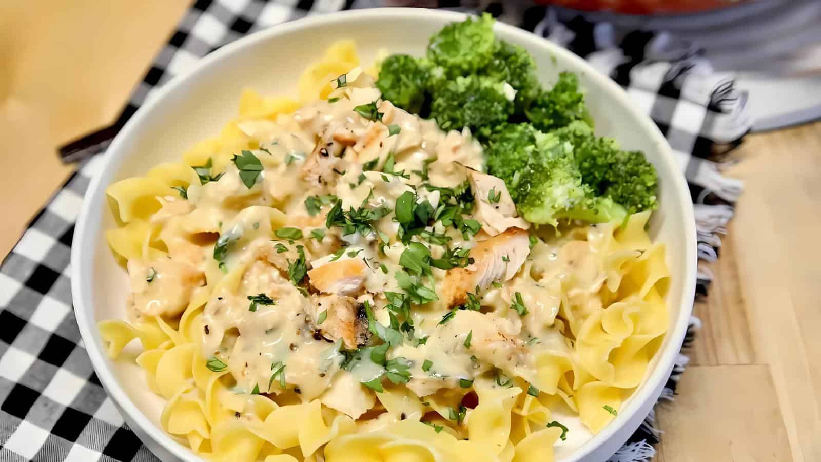 A bowl of pasta topped with a creamy sauce and herbs, accompanied by broccoli, on a black and white checkered napkin.