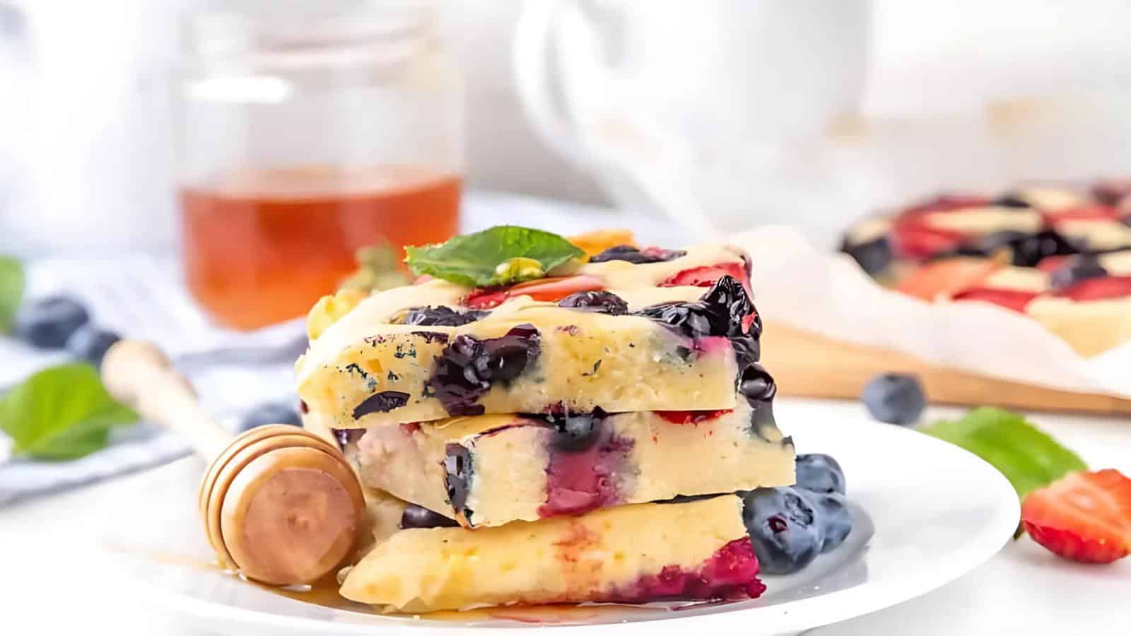A plate with slices of blueberry and cream cheese breakfast bars, garnished with a mint leaf. A honey dipper and jar are in the background.