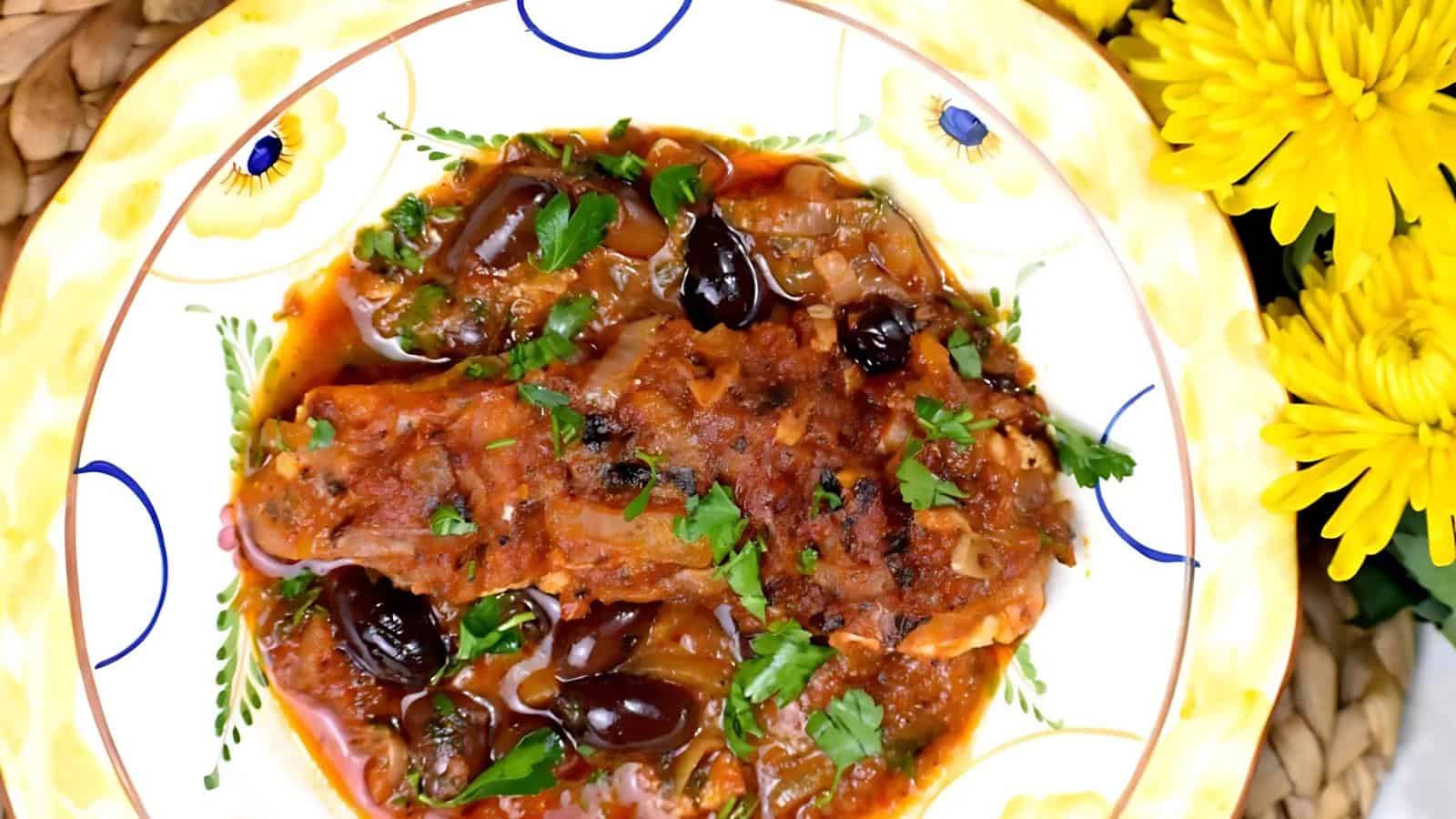 A plate of fish stew with olives and cilantro garnish, next to yellow flowers.