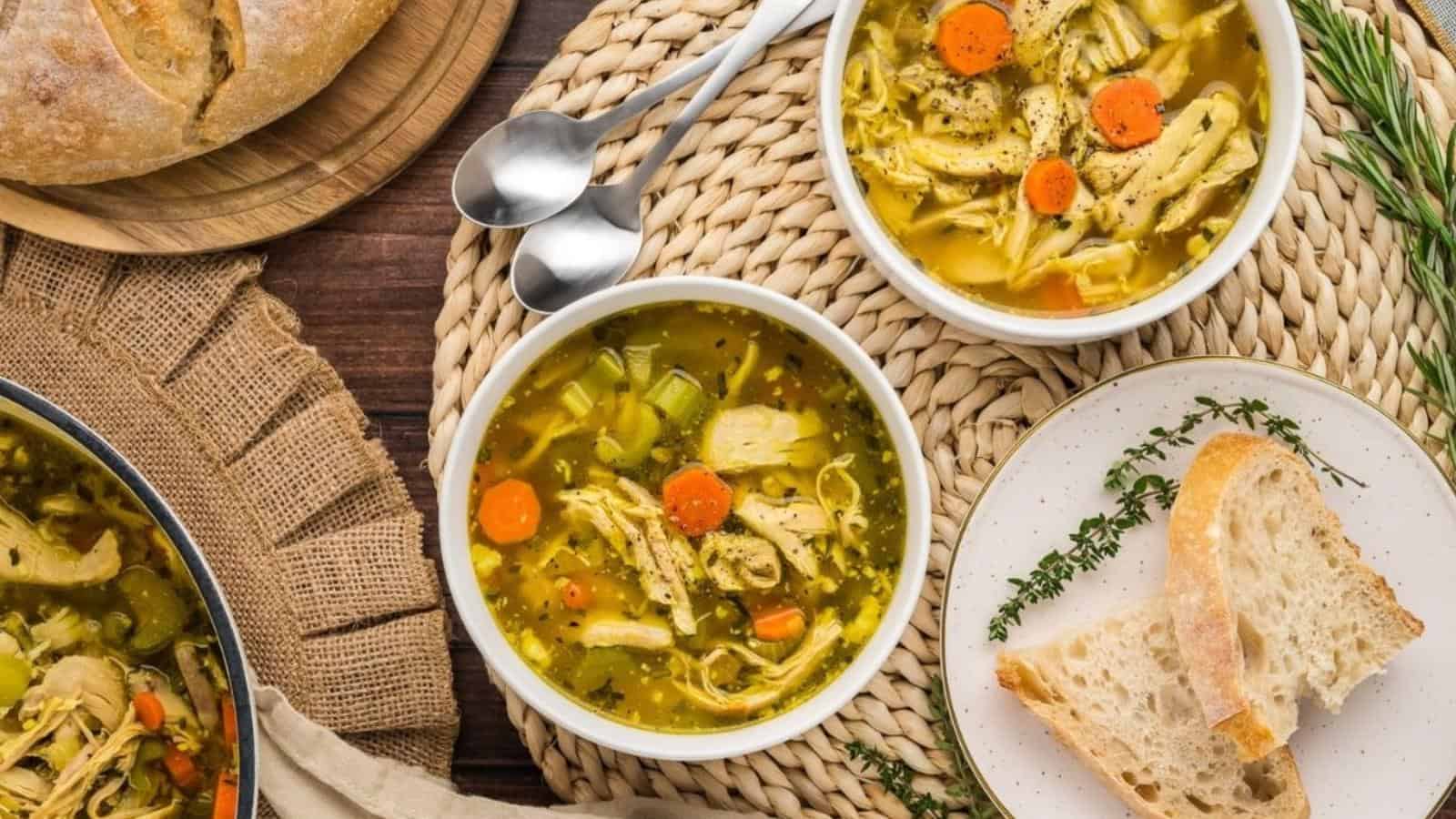 Two bowls of chicken soup with carrots and herbs on a woven mat, accompanied by slices of bread on a plate. Two spoons are placed nearby.
