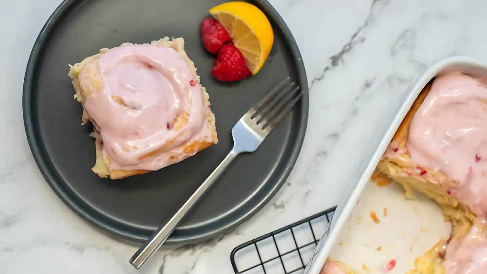 A cinnamon roll with pink icing on a gray plate with raspberries and a lemon slice, next to a partly empty baking dish on a marble surface. A fork is placed on the plate.
