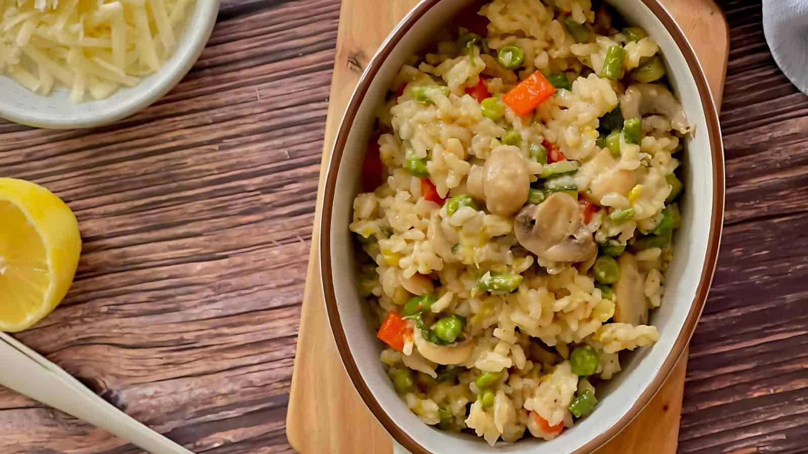 A bowl of risotto with mushrooms, peas, and carrots on a wooden surface, accompanied by a lemon wedge and grated cheese in a small dish.
