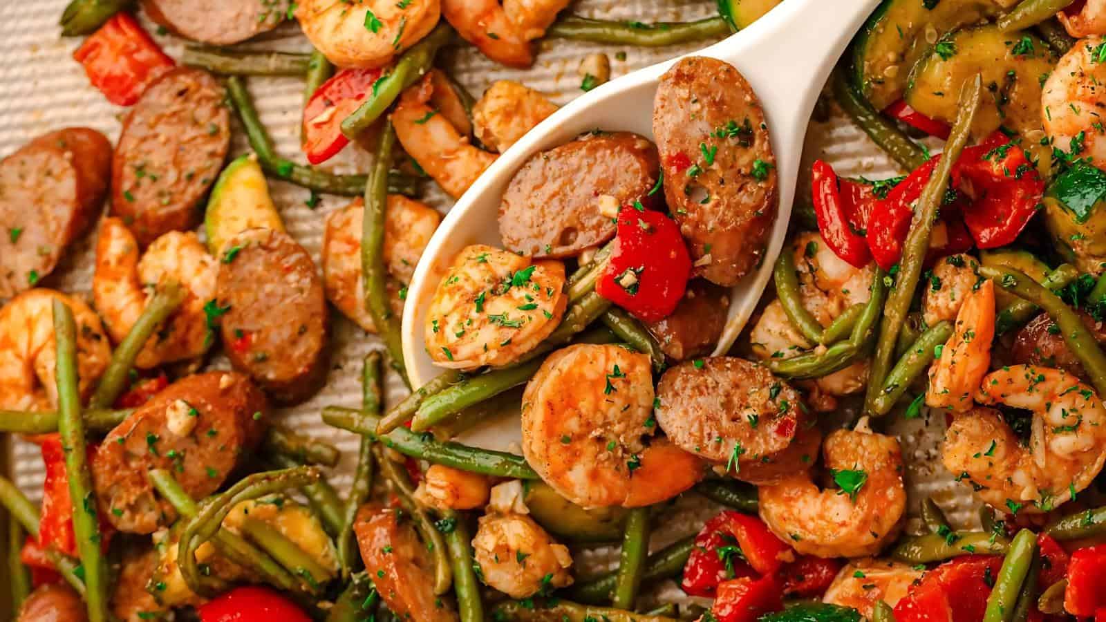 A dish featuring sliced sausage, shrimp, green beans, and red bell peppers, garnished with herbs. A white spoon is scooping a portion from the tray.