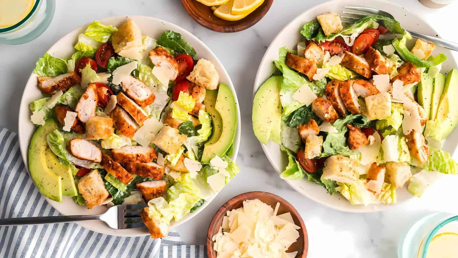 Two plates of Caesar salad with grilled chicken, avocado slices, croutons, cherry tomatoes, and Parmesan cheese. A bowl of lemon wedges and a dish of extra Parmesan are nearby.