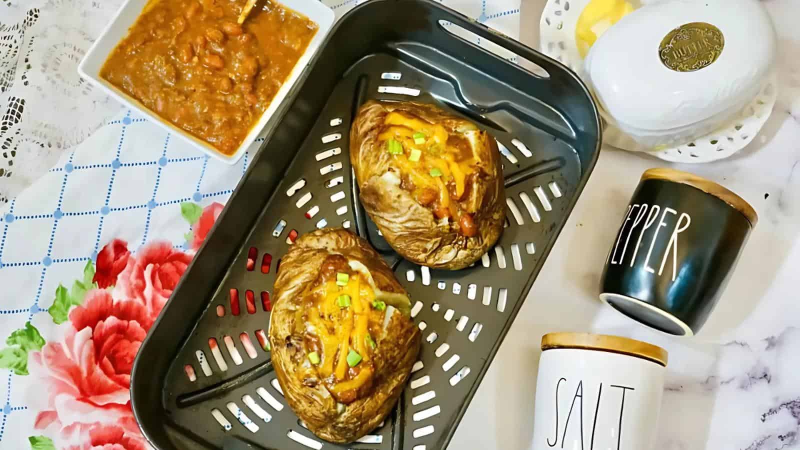 Two baked potatoes topped with cheese and scallions in a baking tray, accompanied by a dish of beans, butter dish, and containers labeled "salt" and "pepper" on a decorative tablecloth.