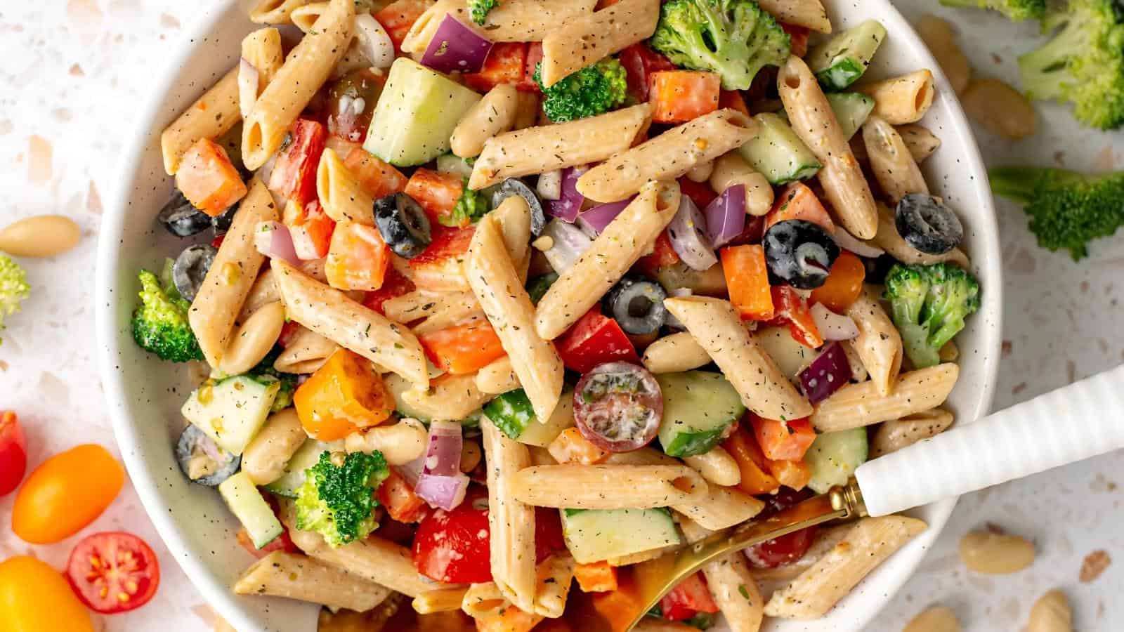 Bowl of pasta salad with penne, broccoli, cherry tomatoes, cucumber, olives, red onion, and beans, mixed with a dressing.
