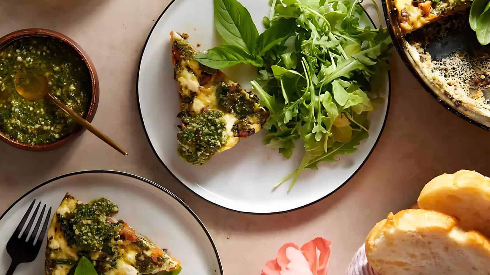Plates of pesto-topped pizza with fresh basil and arugula salad on a table, alongside a bowl of pesto and slices of bread.