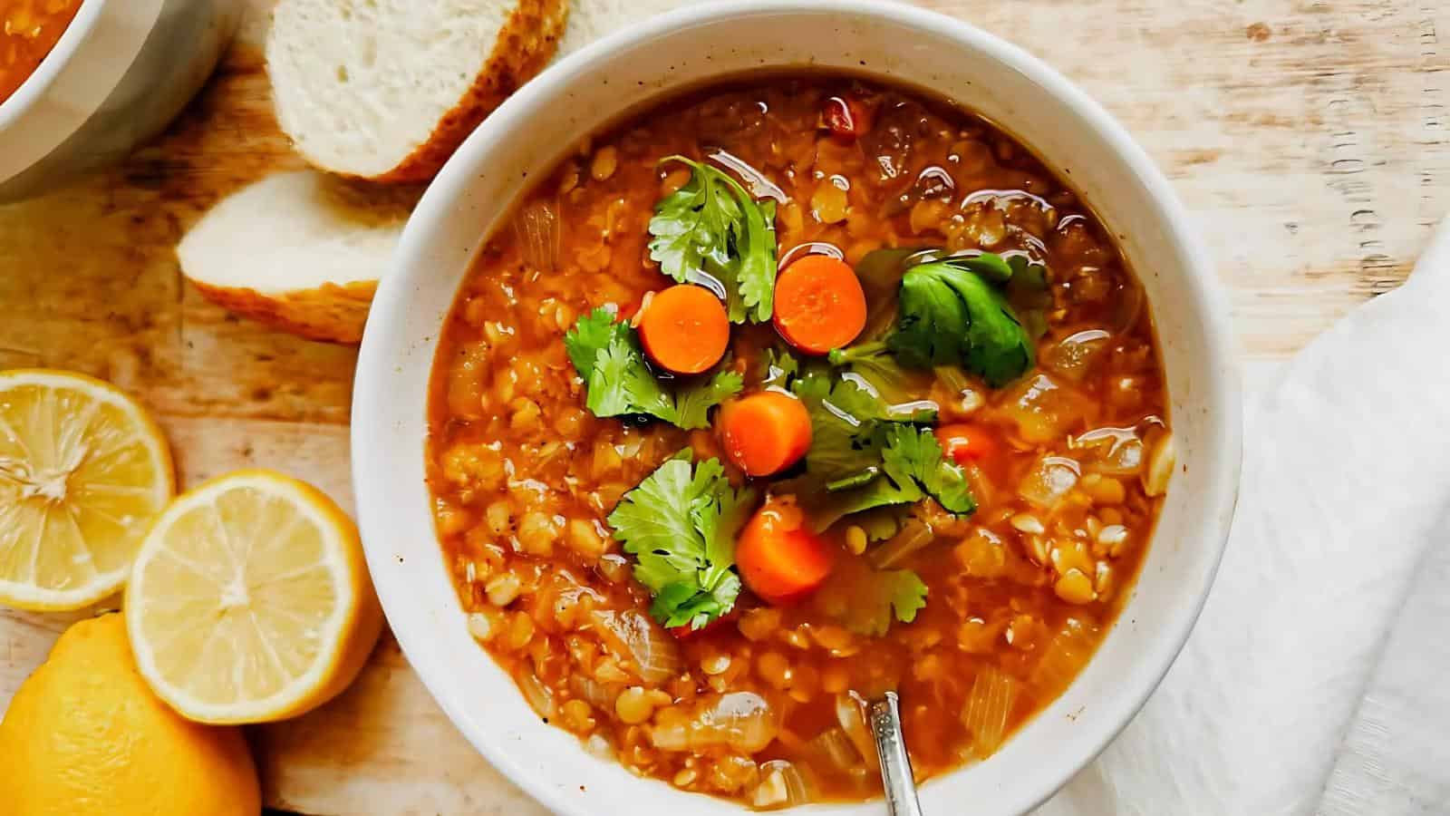 A bowl of lentil soup garnished with cilantro and carrot slices, accompanied by lemon halves and sliced bread on the side.