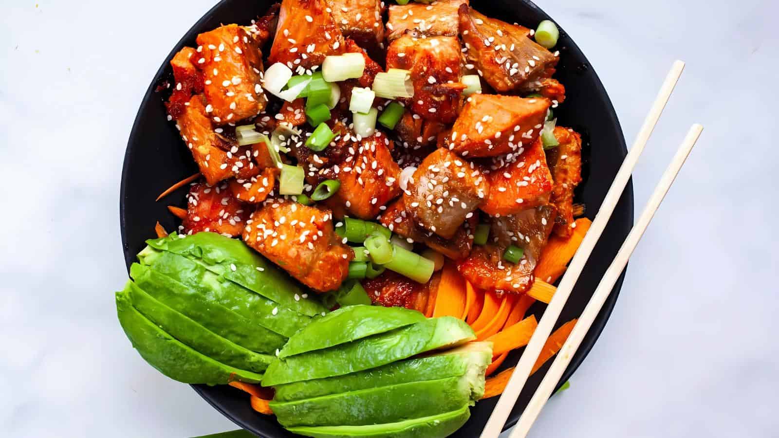 A bowl of tofu glazed with sauce, topped with green onions and sesame seeds, served with sliced avocado and carrot ribbons. Chopsticks rest on the bowl's edge.