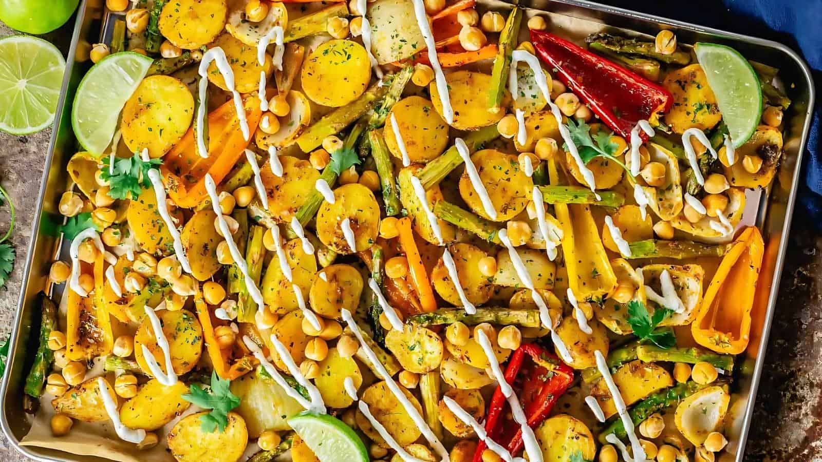 A baking tray with roasted vegetables, including potatoes, bell peppers, and chickpeas, topped with a white drizzle and garnished with lime slices and fresh herbs.