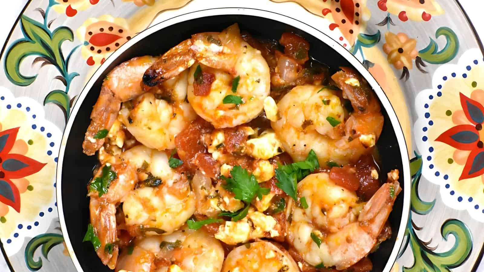 A bowl of cooked shrimp served in a spicy tomato sauce with herbs and cheese, placed on a decorative plate with floral patterns.