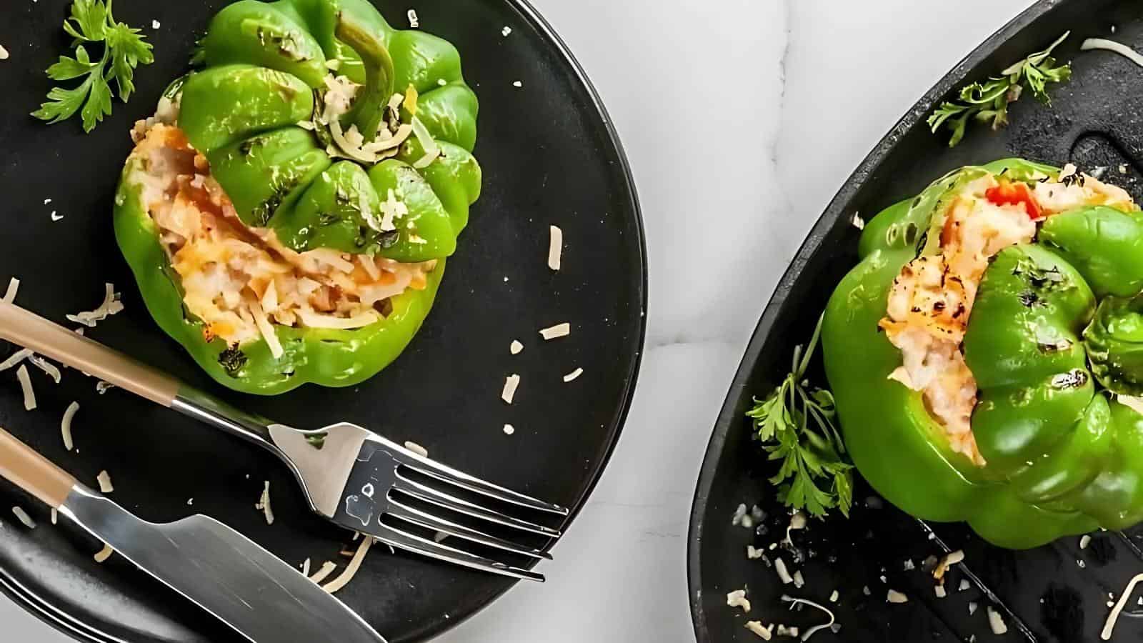 Two black plates with stuffed green bell peppers, filled with cheese and various fillings. A fork and knife are on one plate, and some parsley garnishes the setup.