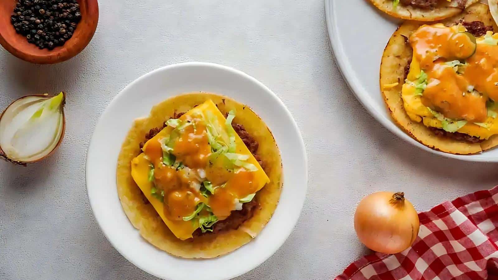 A plate with a tortilla topped with cheese, egg, lettuce, and sauce. Nearby are an onion half, whole onion, pepper bowl, and a red checkered cloth. Another plate is partially visible.
