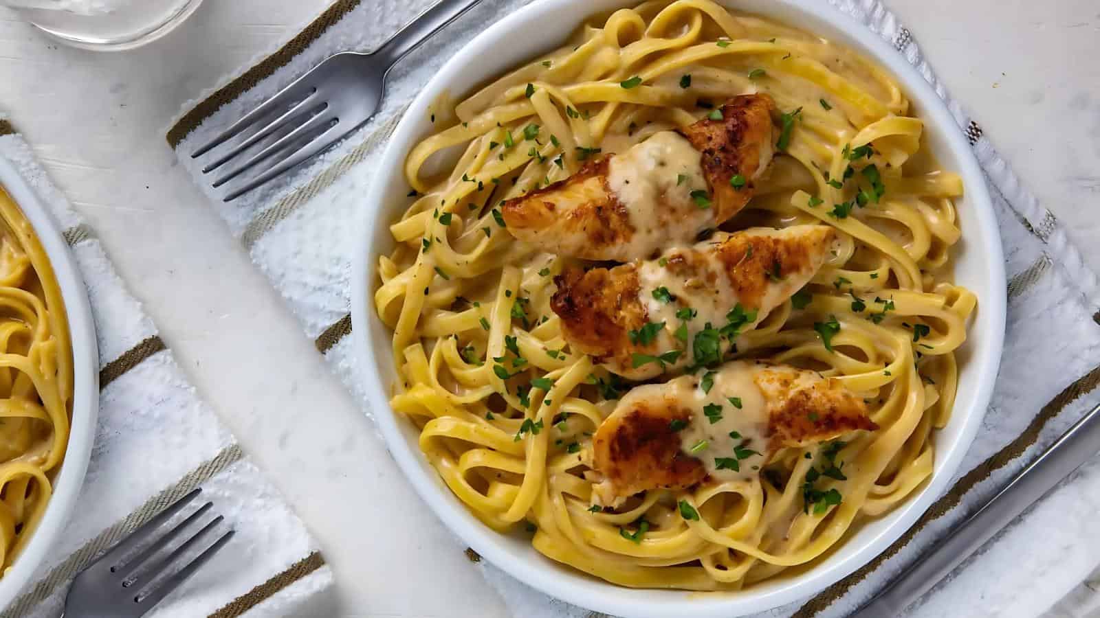 A bowl of fettuccine Alfredo topped with grilled chicken pieces and garnished with parsley, placed on a white cloth with a fork beside it.