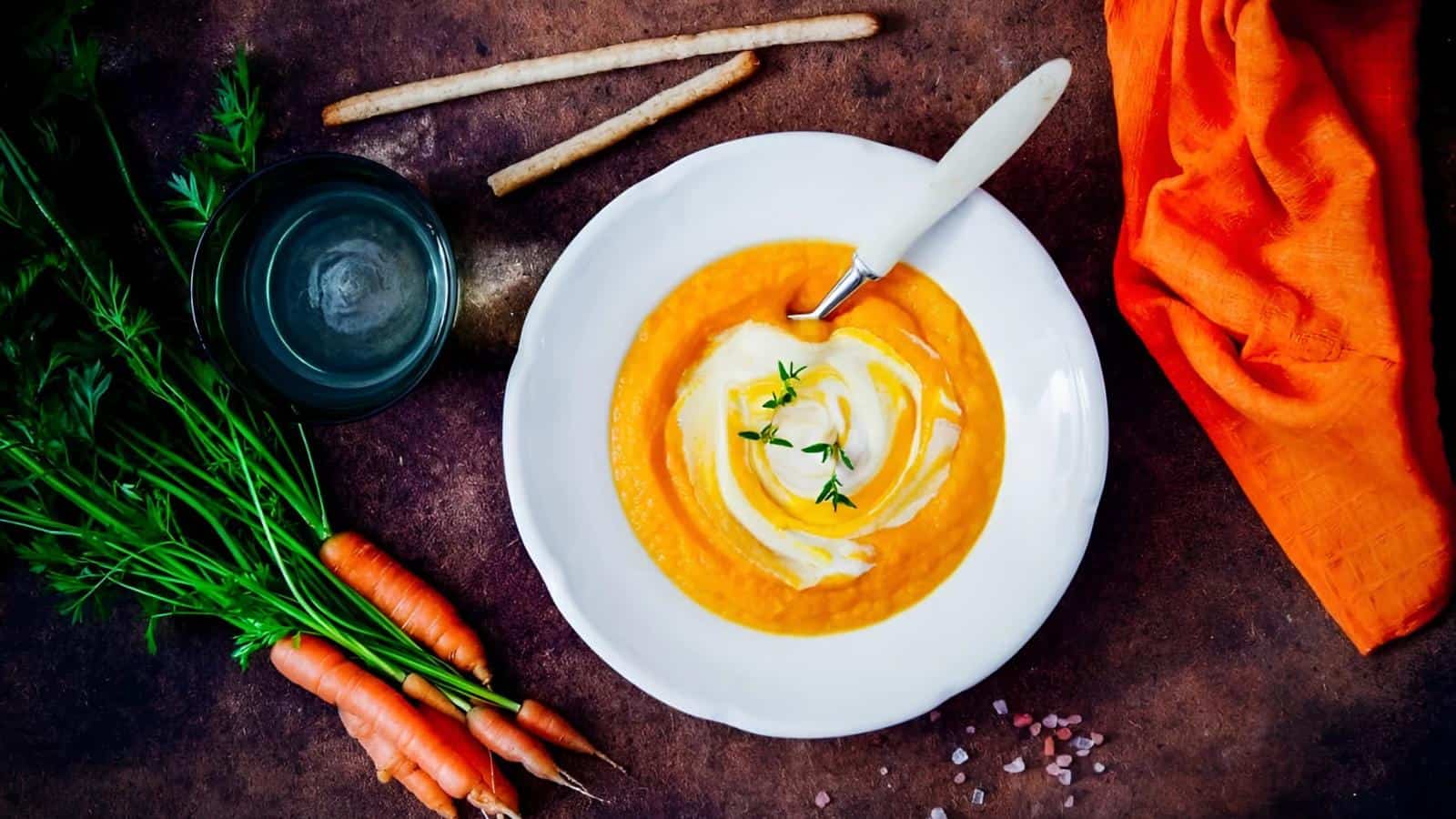 A bowl of carrot soup with cream and herbs on top, surrounded by fresh carrots, breadsticks, a glass of water, and an orange napkin on a dark surface.