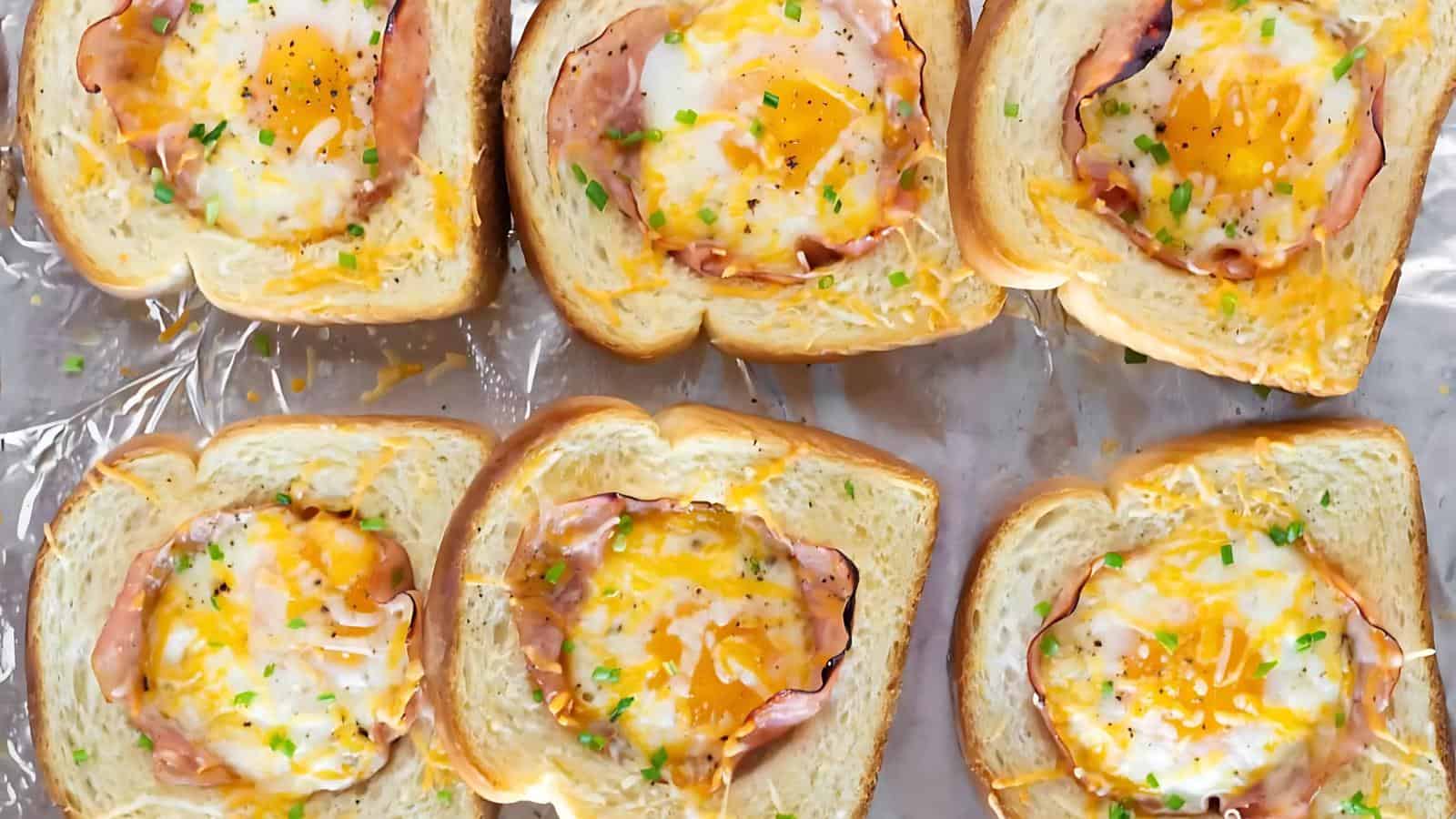 Slices of bread with cooked eggs, cheese, and ham placed in the center, topped with chopped chives, arranged on a baking tray.