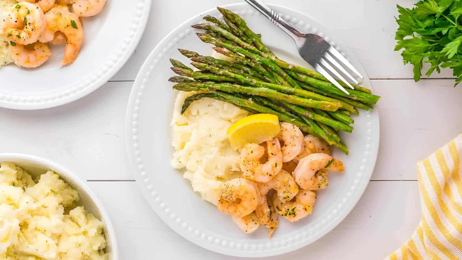 A plate with shrimp, asparagus, mashed potatoes, and a lemon wedge on a white table. Fork on the side, with partially visible mashed potatoes and parsley nearby.