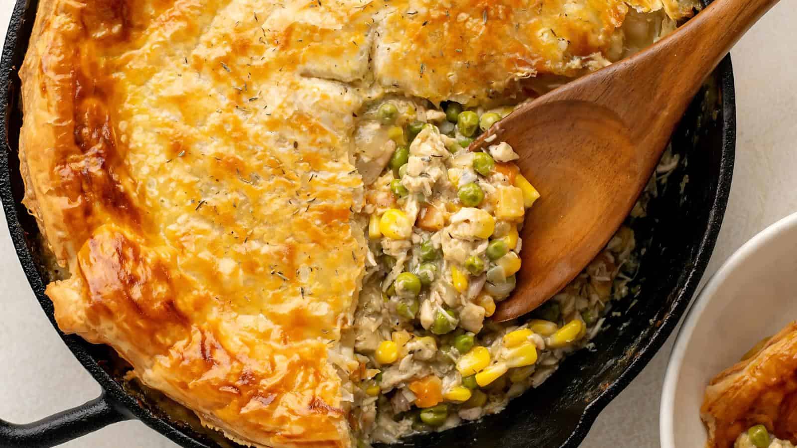 A partially sliced chicken pot pie in a skillet with a wooden spoon, revealing its filling of chicken, corn, peas, and carrots.