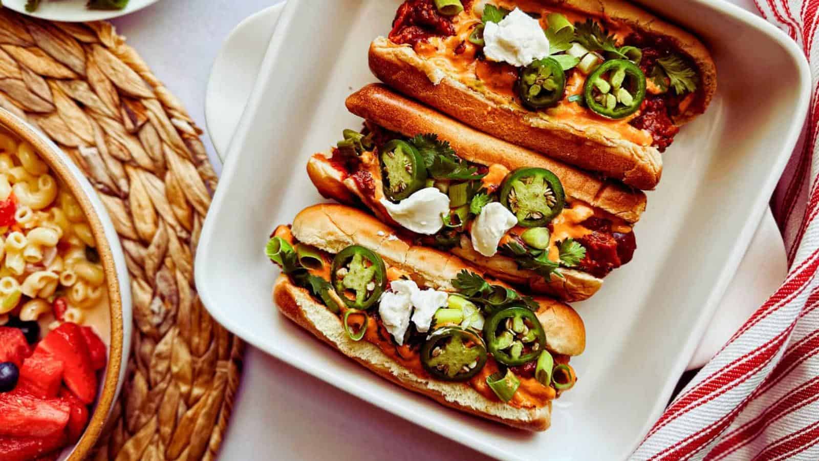 Three hot dogs topped with jalapeños, sour cream, and cilantro in a white dish, with a side dish of macaroni salad visible on the left.