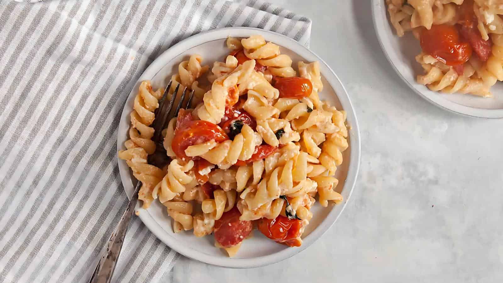 Plate of rotini pasta with cherry tomatoes and cheese on a striped napkin, fork on the side.