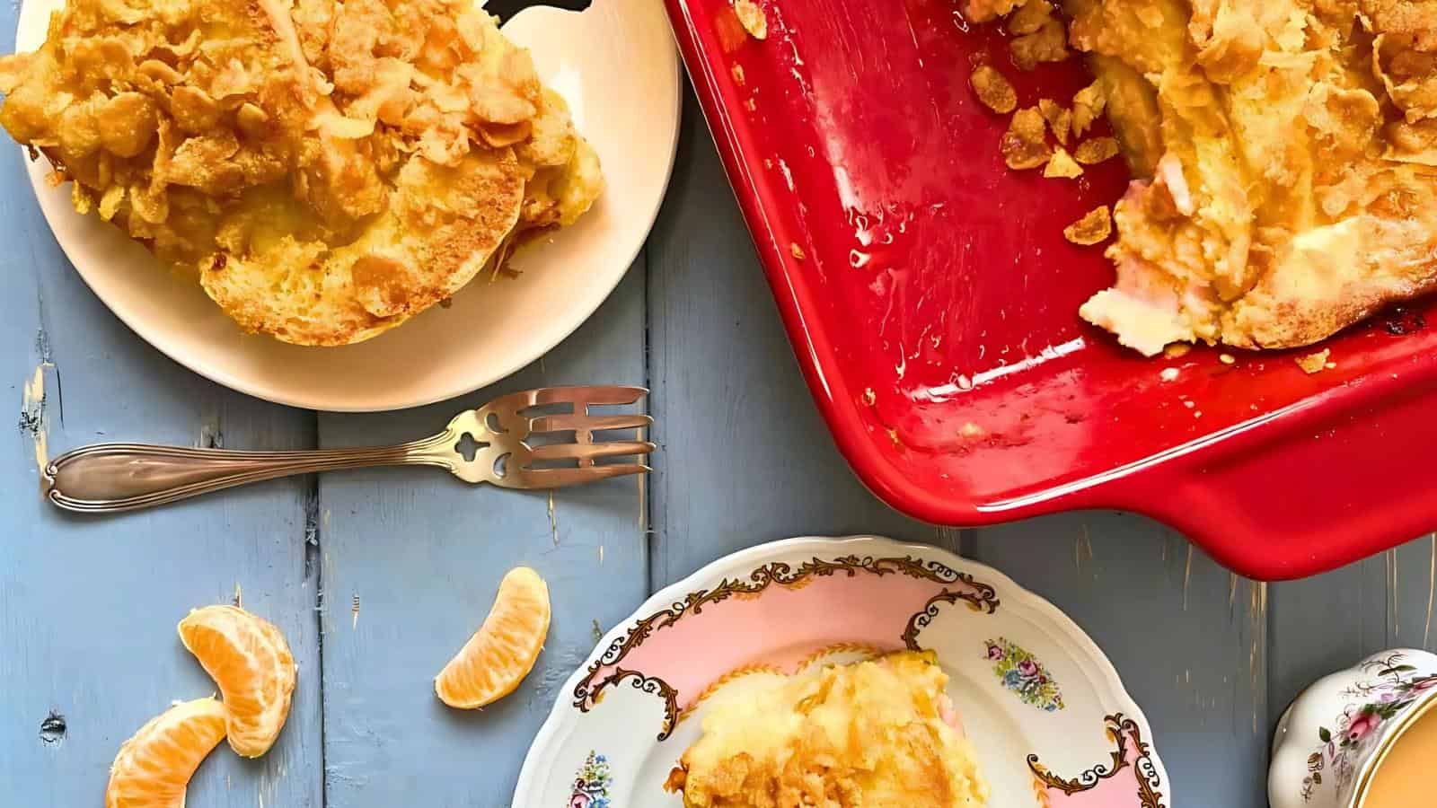 Plates with servings of casserole, a red baking dish with remaining casserole, and a fork. Clementine slices are scattered on a blue wooden table.
