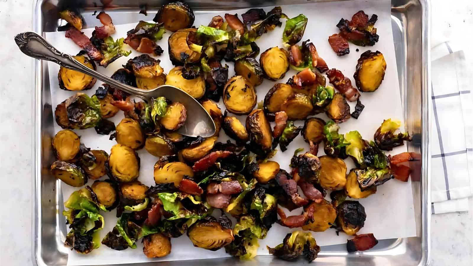 Roasted Brussels sprouts with crispy bacon pieces on a parchment-lined baking tray, with a serving spoon.