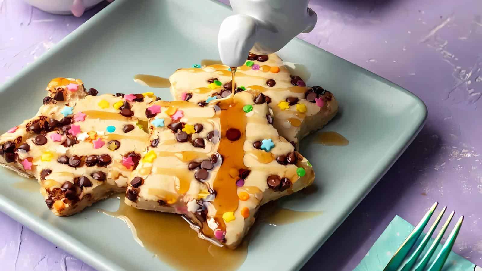 Star-shaped waffles with sprinkles and chocolate chips are being drizzled with syrup on a blue plate.
