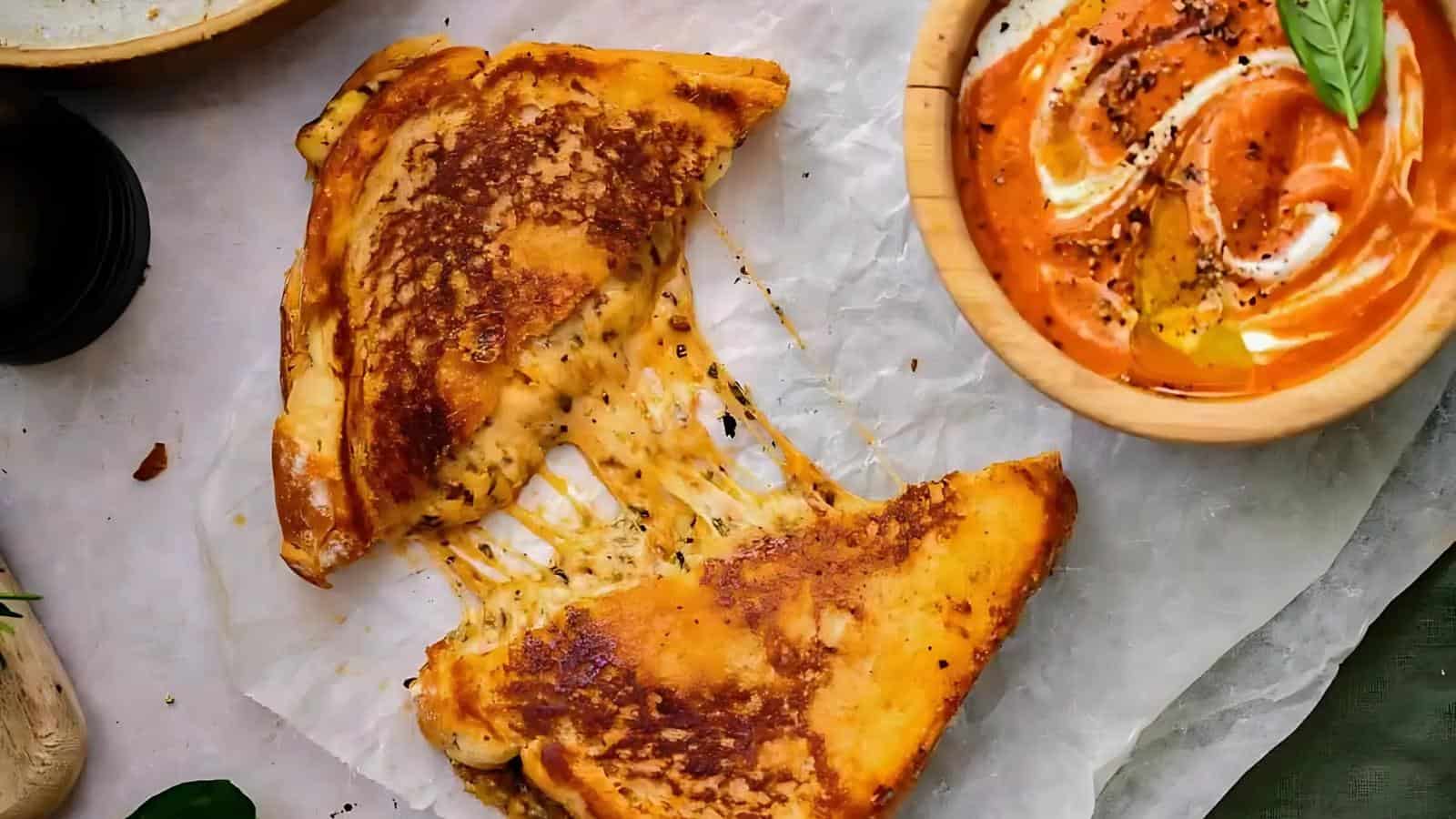 Grilled cheese sandwich with melted cheese pulling apart on parchment paper, next to a bowl of creamy tomato soup.