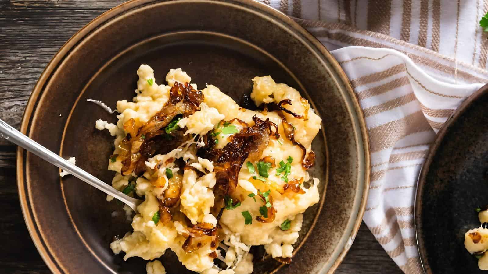 A brown plate with cheesy mashed potatoes topped with caramelized onions and fresh herbs, placed on a wooden table beside a striped cloth.
