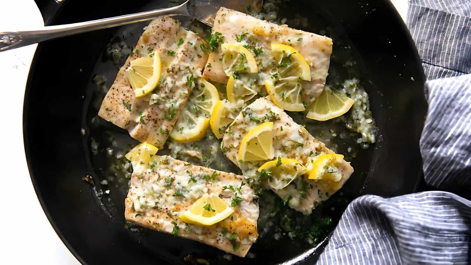 Fish fillets topped with lemon slices and herbs in a black pan, alongside a striped cloth.