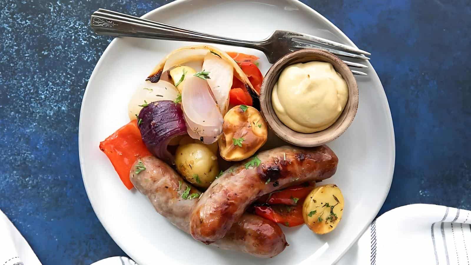 Grilled sausages with roasted red peppers, onions, and potatoes on a white plate, accompanied by a small bowl of mustard and a fork.