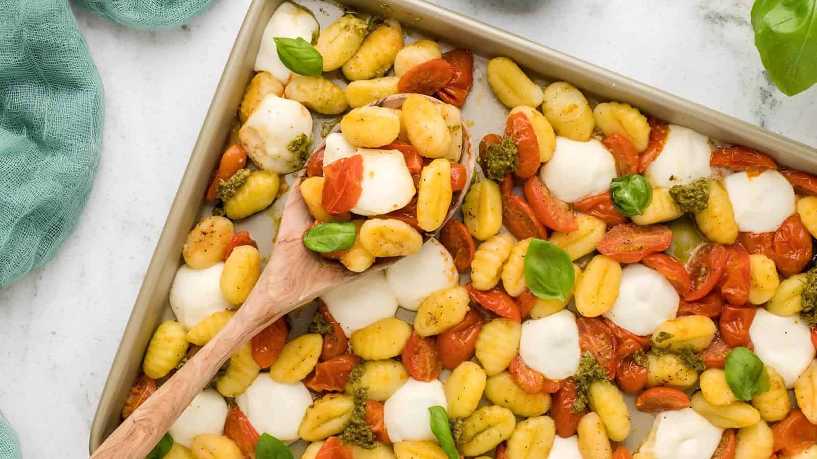 A baking tray of baked gnocchi with cherry tomatoes, melted mozzarella, and basil leaves, with a wooden spoon scooping a portion.