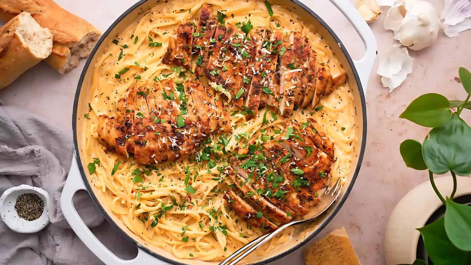 A skillet with creamy pasta topped with three grilled chicken pieces, garnished with chopped parsley. A spoon rests in the dish; bread, pepper, and garlic are visible nearby.