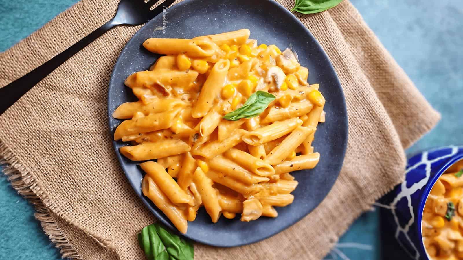 A plate of creamy penne pasta with corn and a basil leaf garnish, placed on a burlap mat with a fork beside it.