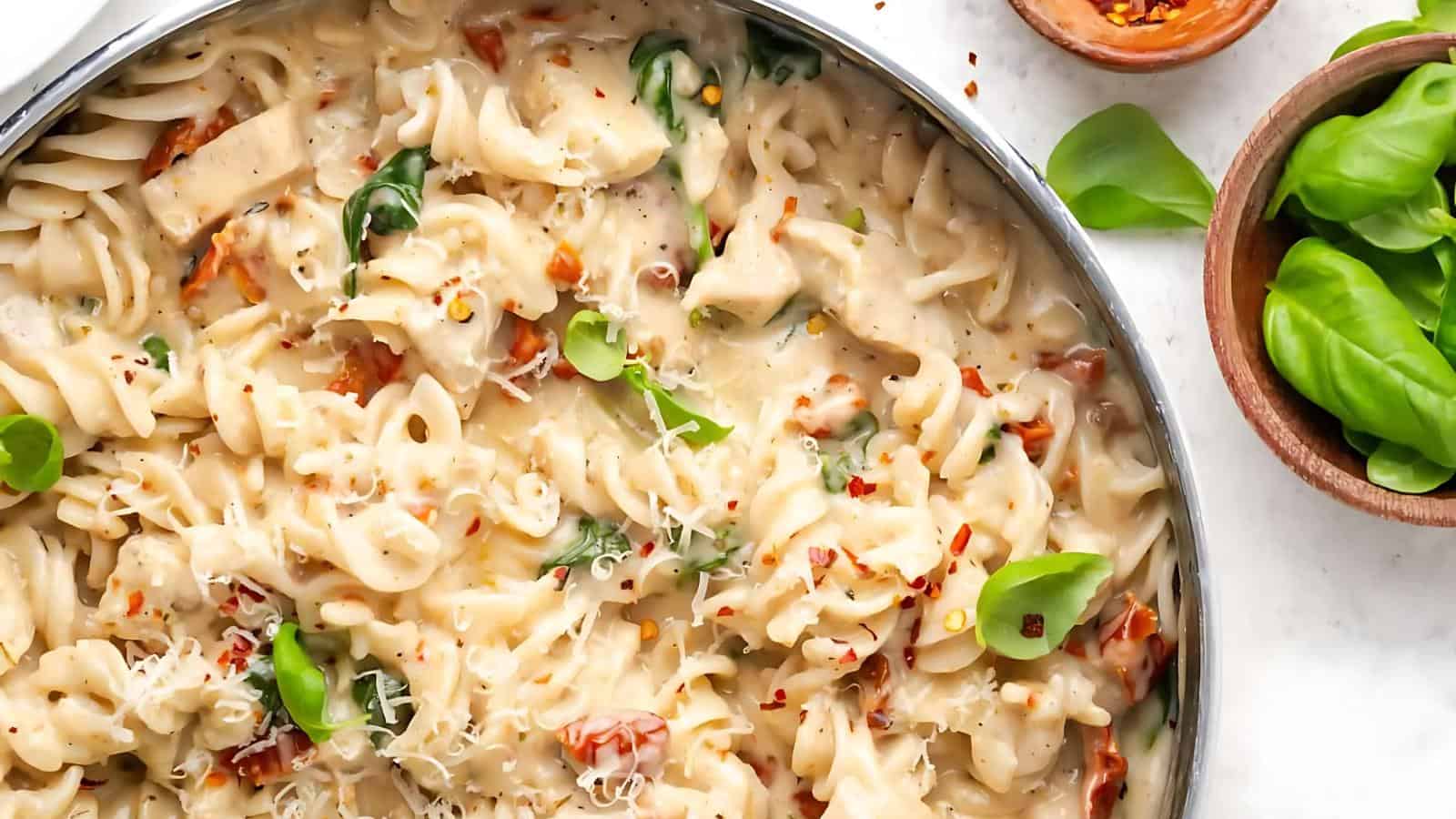 A skillet filled with creamy pasta, garnished with fresh basil leaves and sprinkled with red pepper flakes and grated cheese.
