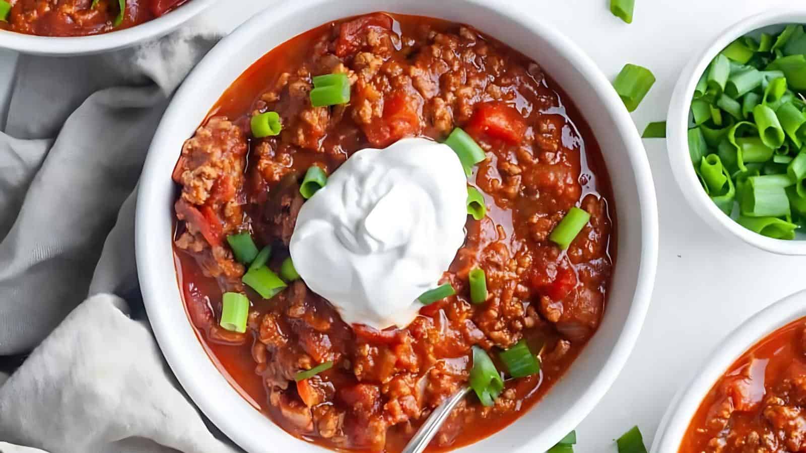 A bowl of chili topped with a dollop of sour cream and garnished with chopped green onions. A cloth napkin and additional green onions in small bowls are nearby.