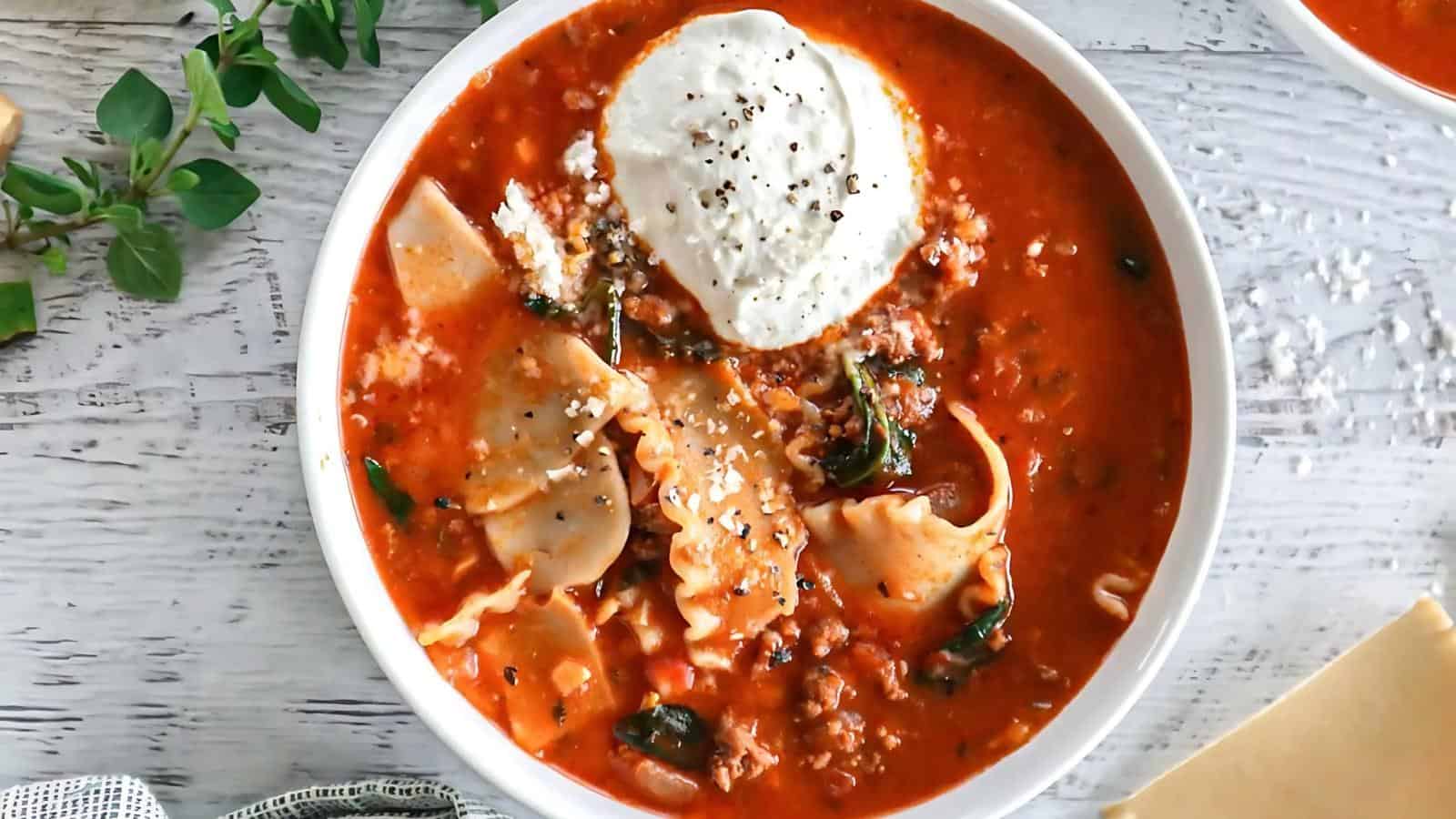A bowl of lasagna soup topped with a dollop of ricotta cheese and black pepper on a light wooden surface, garnished with a sprig of herbs.