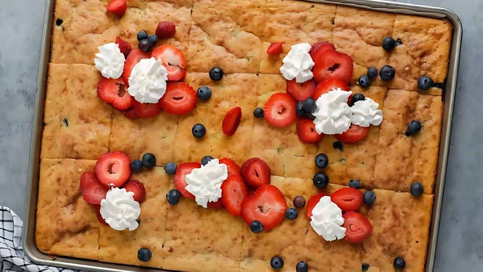 A rectangular sheet cake topped with sliced strawberries, blueberries, and dollops of whipped cream.