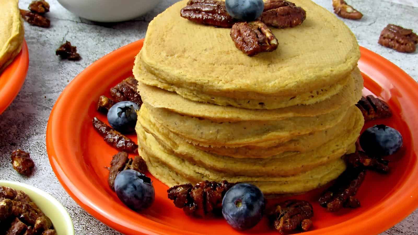 Stack of pancakes on an orange plate, topped with pecans and blueberries. More pecans and blueberries scattered around.