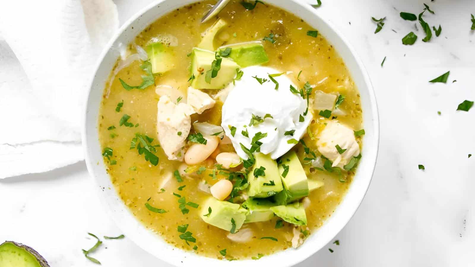 Bowl of chicken chili topped with avocado, white beans, and sour cream, garnished with chopped herbs.