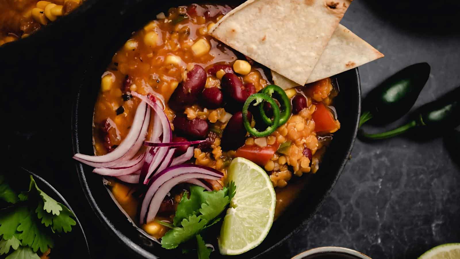 A bowl of chili with kidney beans, corn, and tomato, topped with sliced red onions, jalapeños, cilantro, tortilla chips, and a lime wedge.