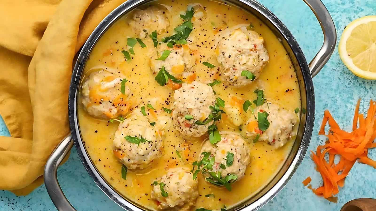 Pot of meatballs in a creamy sauce, garnished with chopped parsley. A yellow cloth and lemon wedge are beside the pot, with shredded carrots nearby on a blue surface.