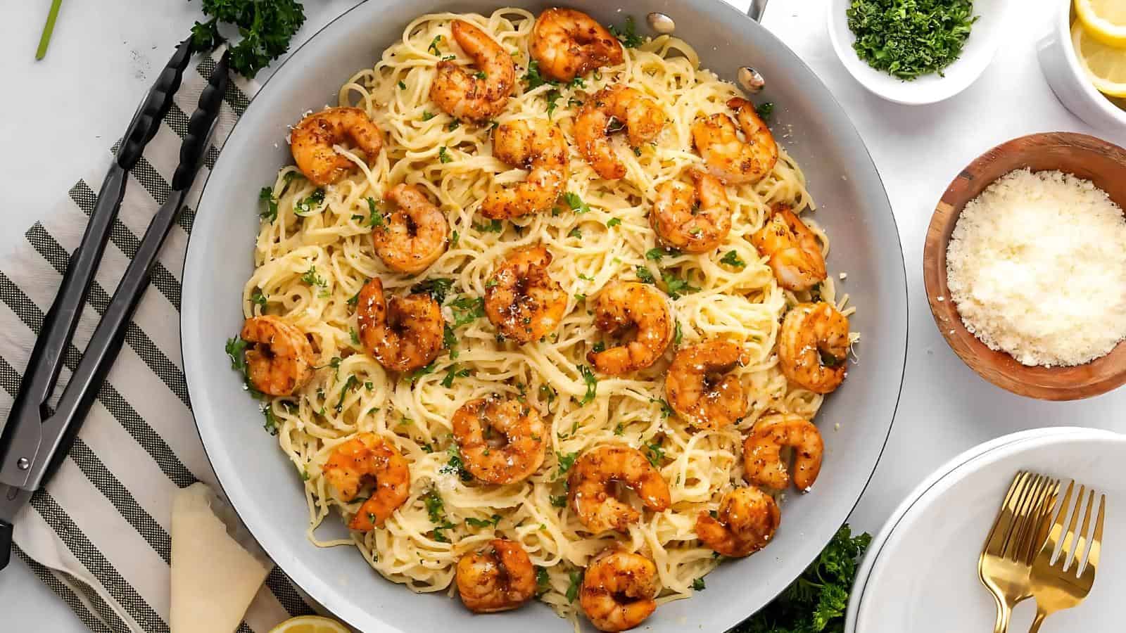 Pan of shrimp scampi with spaghetti, garnished with parsley, next to parmesan cheese, lemon wedges, and tongs on a striped cloth.
