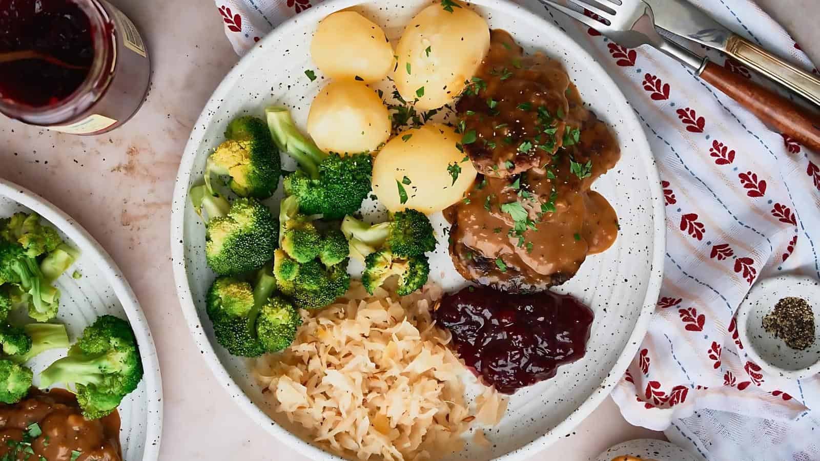 A plate with meatballs in sauce, boiled potatoes, broccoli, sauerkraut, and a dollop of red jam, garnished with herbs.
